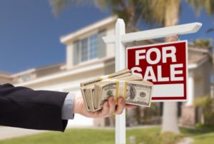 a person holding out a handful of cash in front of a house with a for sale sign in front of it